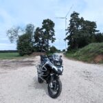 Motorrad (weiße BMW R 1200 GS) steht auf einem Schotterweg. Im Hintergrund sind zwei Windkraftanlagen und Bäume zu sehen.