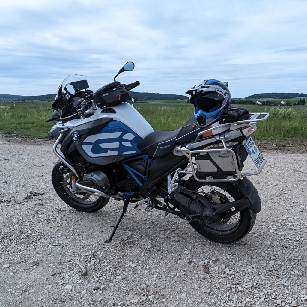 Weiß-blaues Motorrad (BMW R 1200 GS) mit Seitenkofferhalterung und Helm auf einem Schotterweg in hügeliger Landschaft.