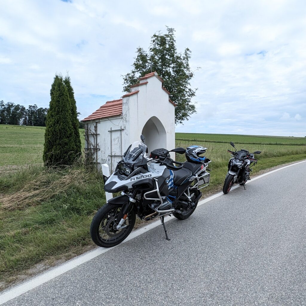 Zwei Motorräder (silberne BMW R 1200 GS und schwarzes Naked Bike) parken vor einer weißen Feldkapelle am Straßenrand.