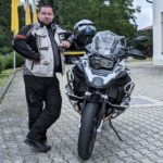 Mann in Motorradbekleidung (grau-schwarz, Textil) lehnt an einer weißen BMW R 1200 GS. Der Helm hängt am Spiegel.