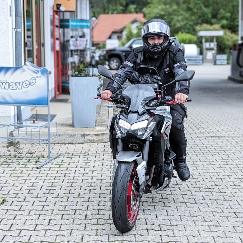 Mann in schwarzer Motorradkleidung und Helm sitzt auf einem schwarzen-silbernen Kawasaki Z900 Motorrad mit roten Felgen auf einem gepflasterten Vorplatz.