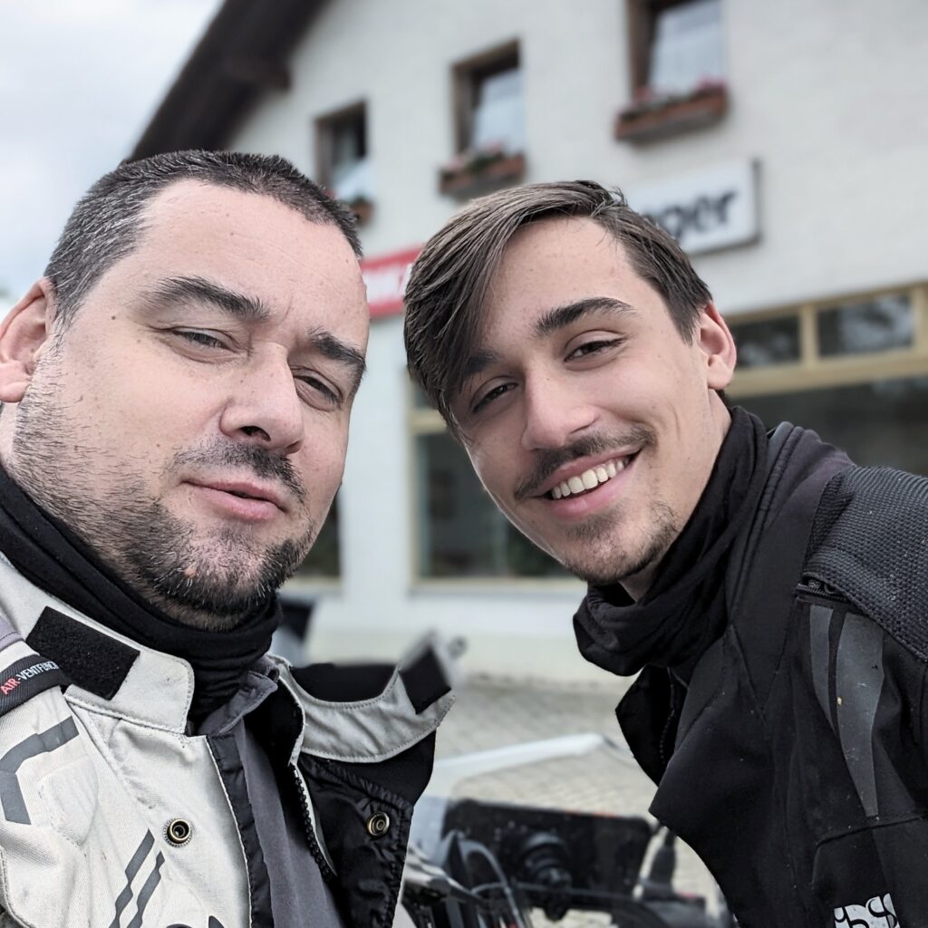 Zwei lächelnde Männer in Motorradkleidung posieren für ein Selfie vor einem Gebäude im Hintergrund.