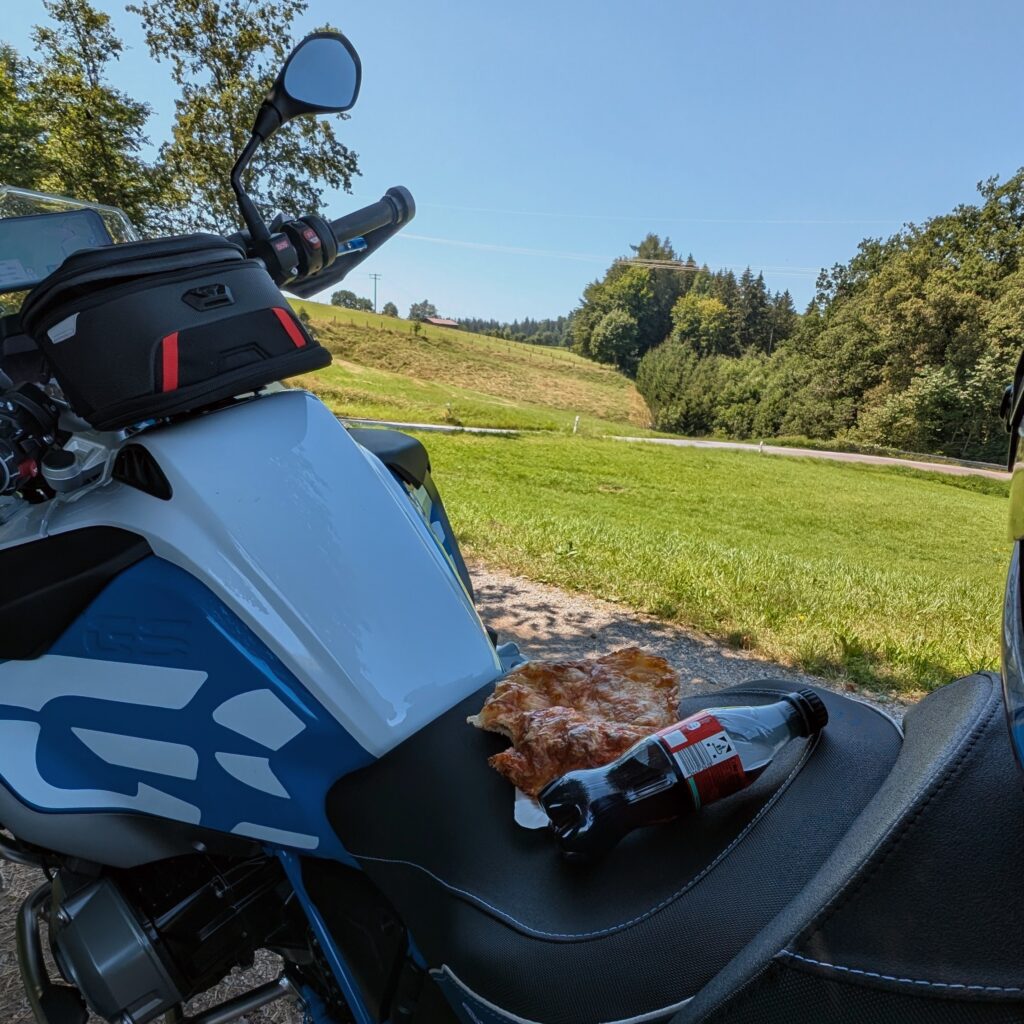 Motorrad (BMW R1200GS) mit blau-weißer Lackierung und einem Stück Pizza und einer Coca-Cola Flasche auf dem Sitz.