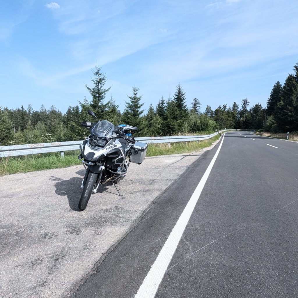 Eine schwarze BMW R 1200 GS Adventure mit Gepäck auf einem sonnigen Landstraßenstreifen am Waldrand.