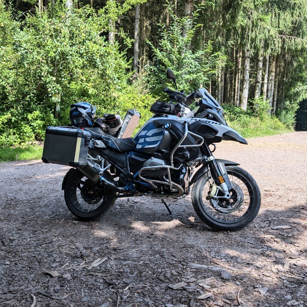 Graues BMW-Motorrad mit Packtaschen und Sturzbügeln steht auf einem Schotterweg im Wald.