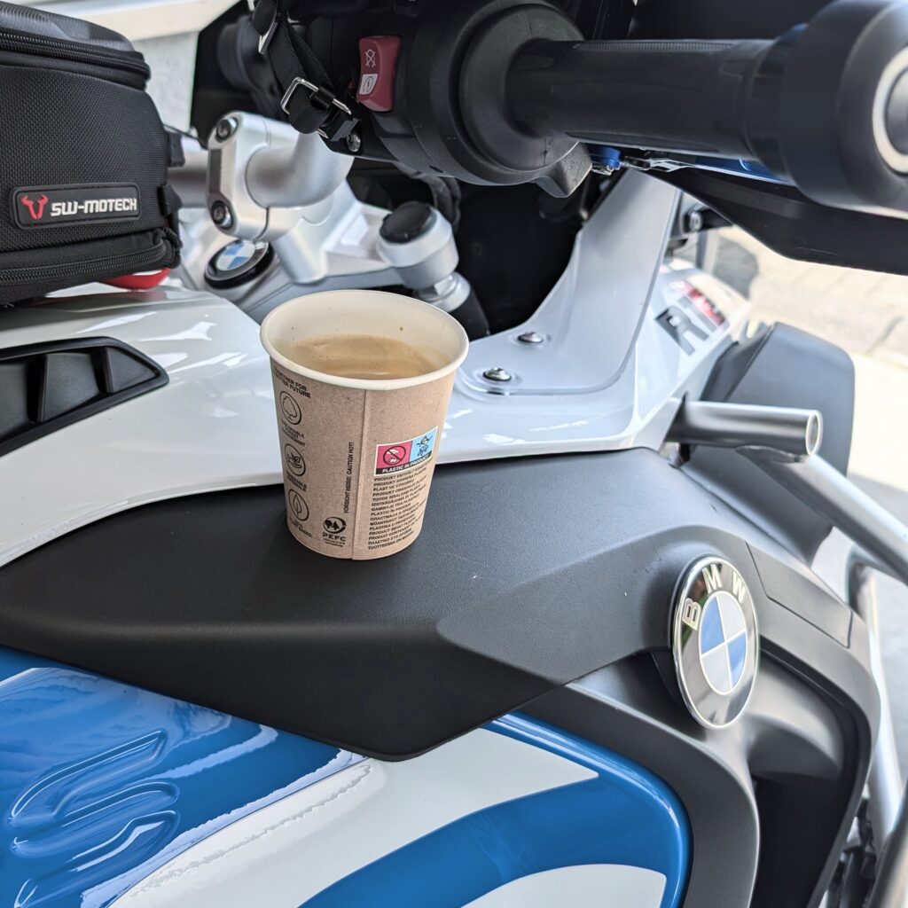 Kaffee im Einwegbecher aus braunem Karton steht auf dem schwarzen Cockpit einer weiß-blauen BMW-Motorradverkleidung.
