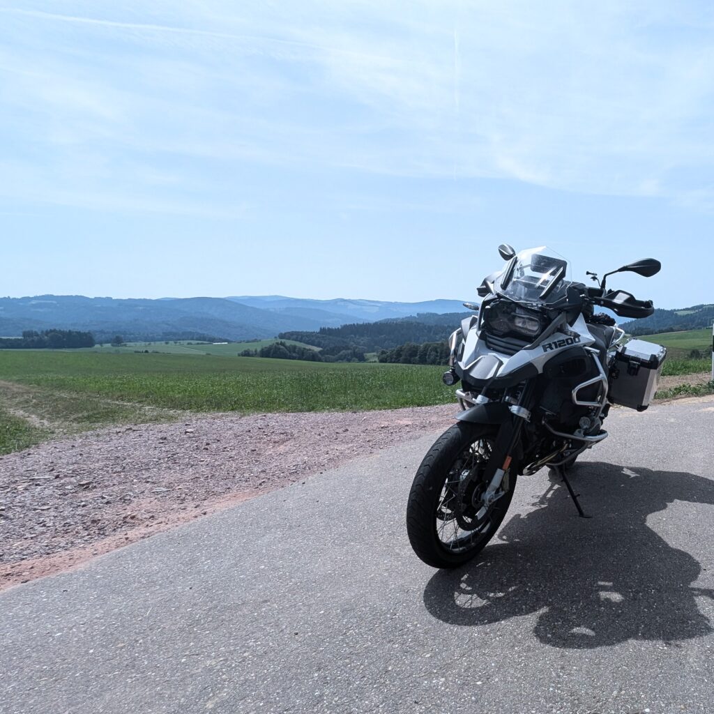 Grau-weißes BMW R 1200 Adventure Motorrad mit Seitenkoffern und Sturzbügel, auf einem Schotterweg vor dichtem Grün.