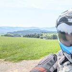 Der behelmte Kopf eines Motorradfahrers mit blauer Visierkante vor einer weiten, sonnigen Hügellandschaft mit grünen Wiesen.