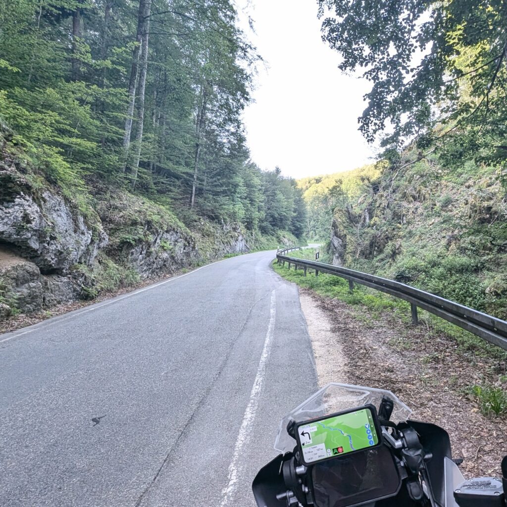 Motorradfahrt in einer Waldschlucht, Sicht auf die kurvige Straße mit Leitplanke und einem Navigationsgerät am Lenker.