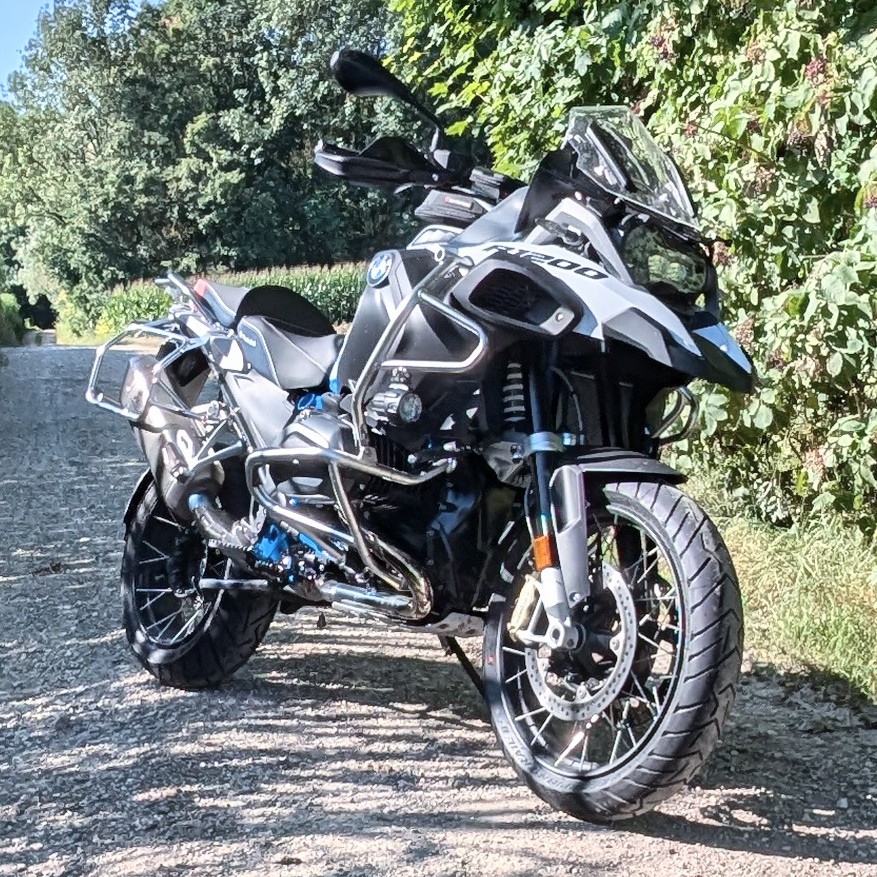 Schwarze und weiße BMW R 1200 GS Adventure mit silbernem Sturzbügel, auf einem Schotterweg mit grünen Büschen im Hintergrund.