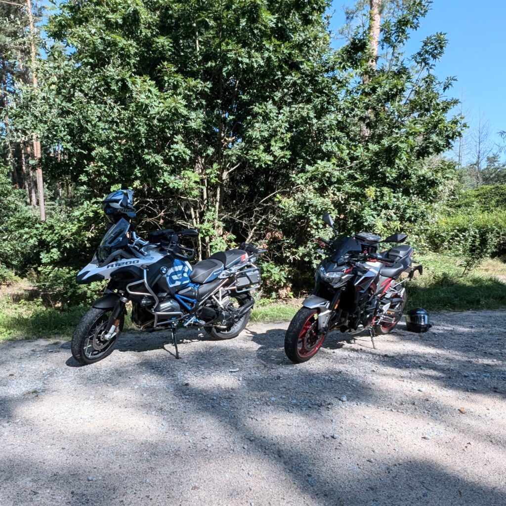 Zwei Motorräder parken auf einem Schotterweg vor einem großen Baum: eine BMW R 1200 GS und eine graue Kawasaki Z900.