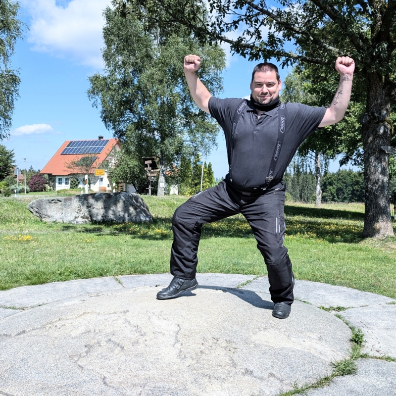 Ein Mann in schwarzer Motorradkleidung (Polo-Shirt, Hose, Hosenträger) steht jubelnd auf einer grauen Steinplatte vor einem Haus mit Solardach.
