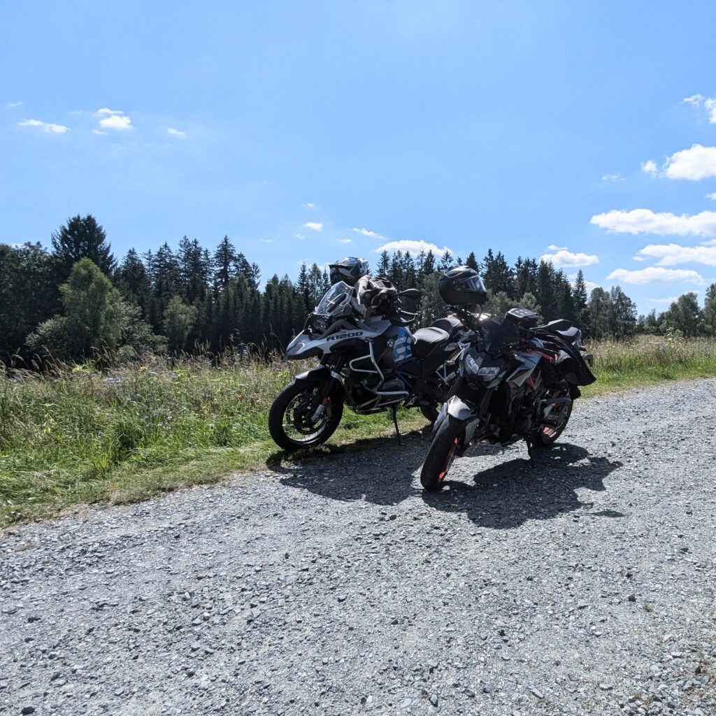 Zwei Motorräder (eine silberne BMW R1200 GS, ein dunkles Sportbike) parken auf einem Schotterweg vor einem Waldstück bei Sonnenschein.