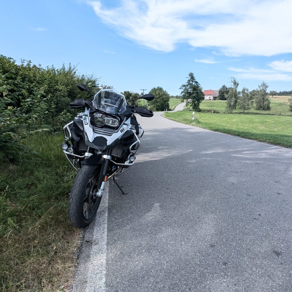 Eine weiße BMW R 1200 GS Adventure ist am Straßenrand in ländlicher Umgebung mit Wiesen und Bauernhof abgestellt.