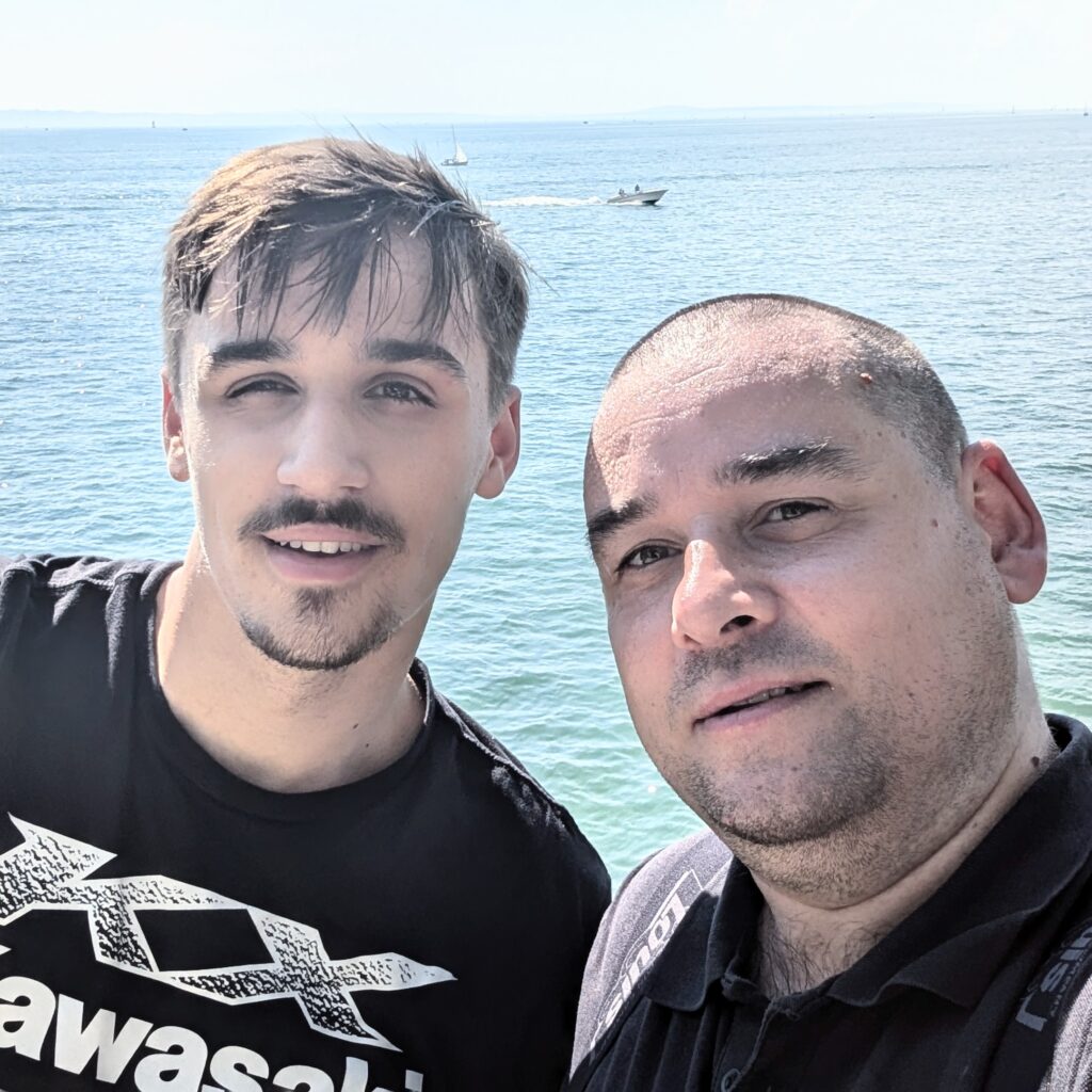 Selfie zweier Männer vor dem Meer, einer trägt ein schwarzes Kawasaki-T-Shirt.