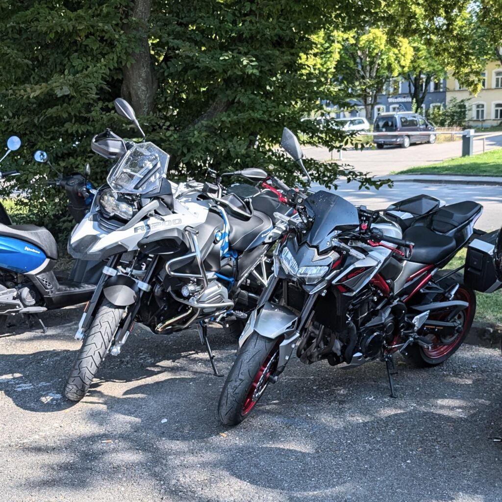 Zwei Motorräder (weiße BMW R1200 GS und silberne Kawasaki Z900) parken auf einem sonnigen Parkplatz.