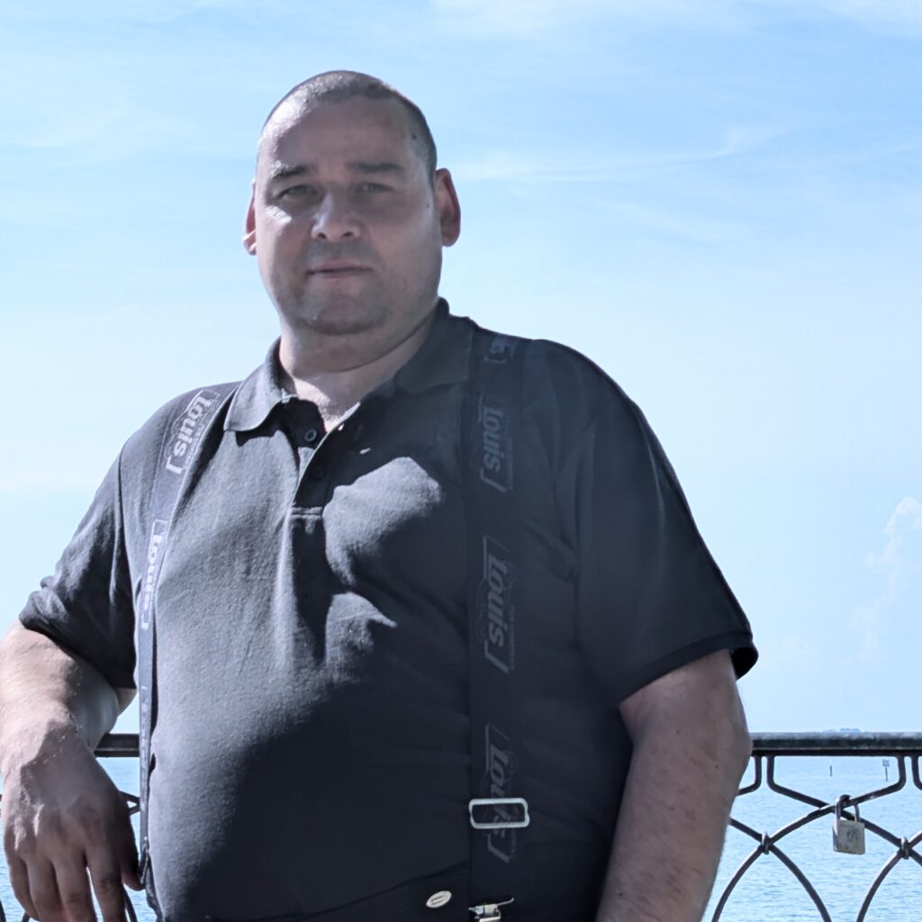 Mann mit schwarzem Louis-Hosenträger und schwarzem Polo-Shirt steht vor einem Metallgeländer am Meer.