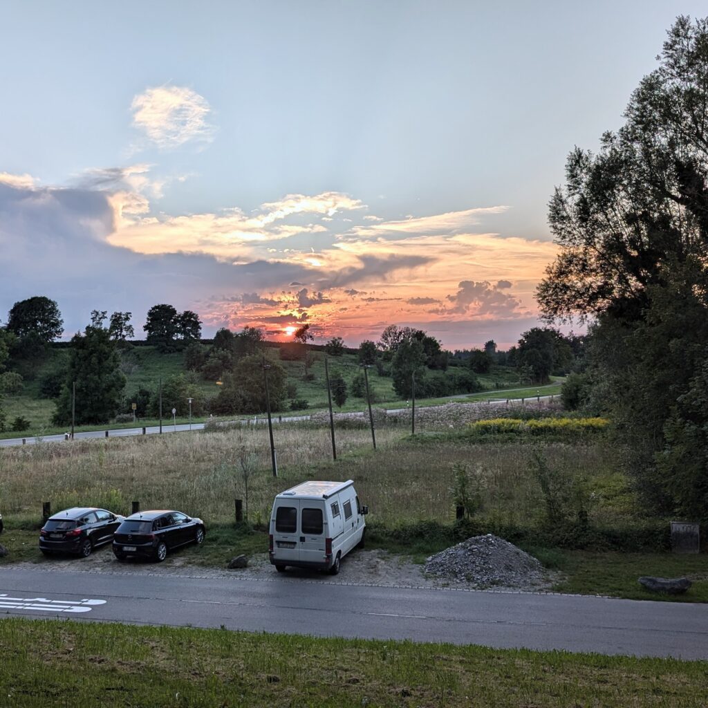 Wohnmobil und zwei PKWs parken vor einem Feld. Im Hintergrund geht die Sonne dramatisch über Hügeln unter.