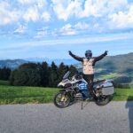 Motorradfahrer mit Helm steht jubelnd auf einer BMW R 1200 GS vor einer hügeligen Waldlandschaft.