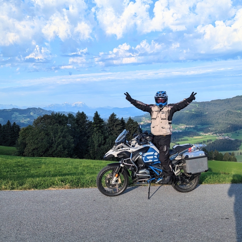 Motorradfahrer mit Helm steht jubelnd auf einer BMW R 1200 GS vor einer hügeligen Waldlandschaft.