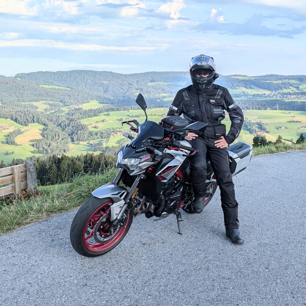 Motorradfahrer sitzt auf einer silbernen Kawasaki Z900 mit roter Felge vor einer grünen Hügellandschaft.