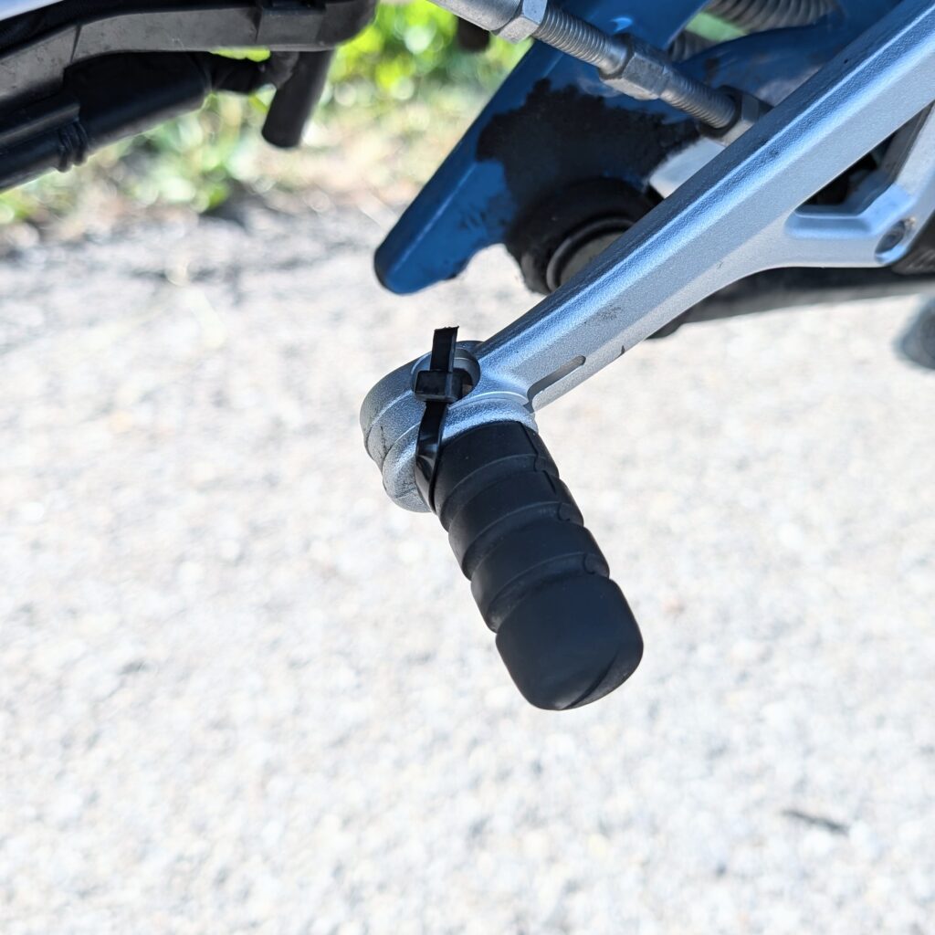 Detailaufnahme des silbernen Schalthebels eines Motorrads mit schwarzem Gummi und einem Kabelbinder.