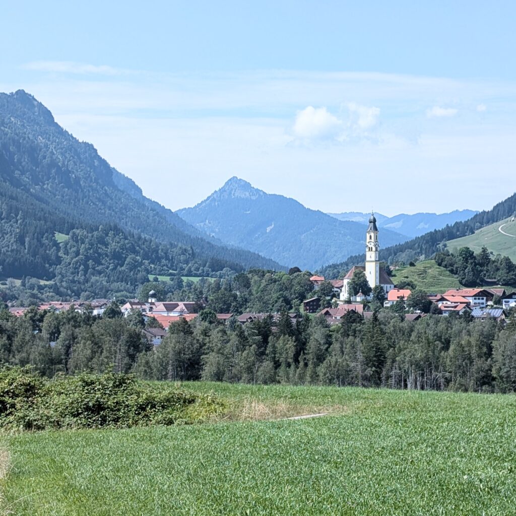 Panoramablick auf ein bayerisches Alpendorf mit einer weißen Kirche vor einem Hintergrund aus grünen Berghängen.