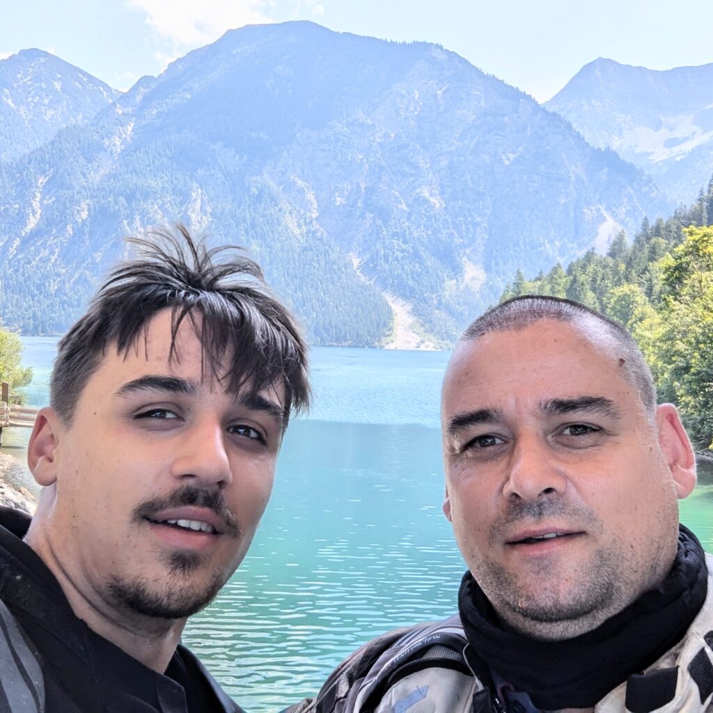 Selfie zweier Motorradfahrer vor einem türkisfarbenen Bergsee und hohen bewaldeten Alpengipfeln.