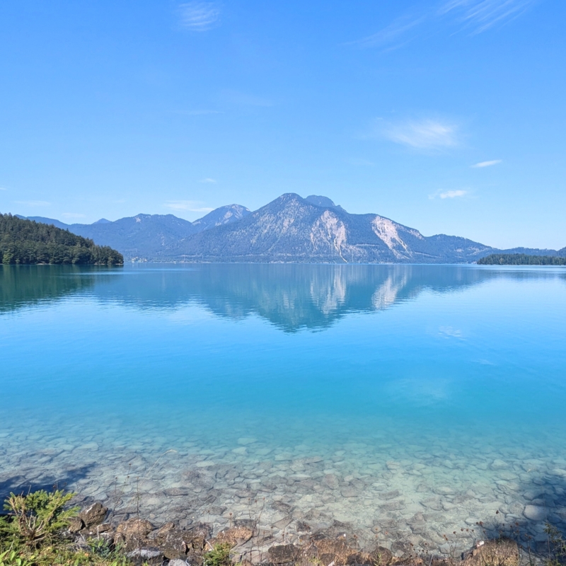 Blau-türkiser Bergsee mit klarem Wasser und Steinen am Ufer. Im Hintergrund spiegeln sich bewaldete Berge.