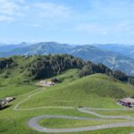 Eine schmale asphaltierte Serpentinenstraße windet sich durch sanfte, grüne Berghügel in einer weiten Landschaft.
