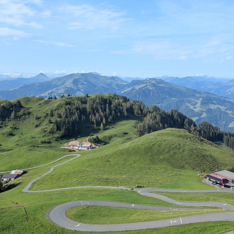 Eine schmale asphaltierte Serpentinenstraße windet sich durch sanfte, grüne Berghügel in einer weiten Landschaft.