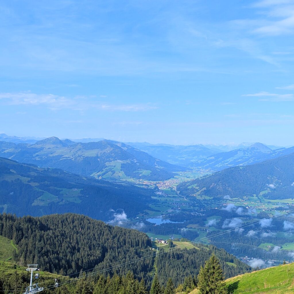 Panorama-Blick über ein weites, grünes Bergtal mit einem kleinen See, einem Dorf und einer Seilbahnstation.