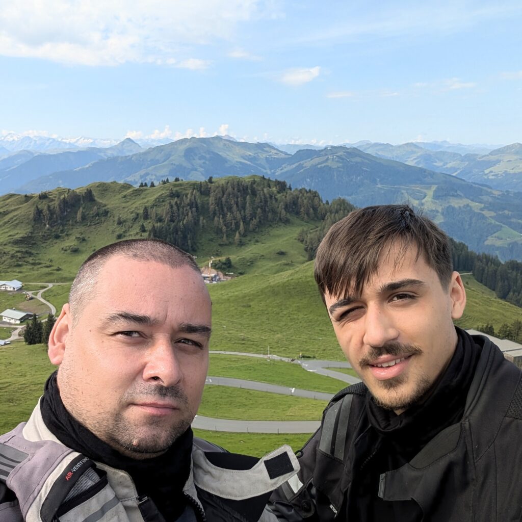 Selfie von zwei Motorradfahrern in Schutzkleidung vor einer weiten Berglandschaft mit grünen Hängen und Serpentinenstraße.