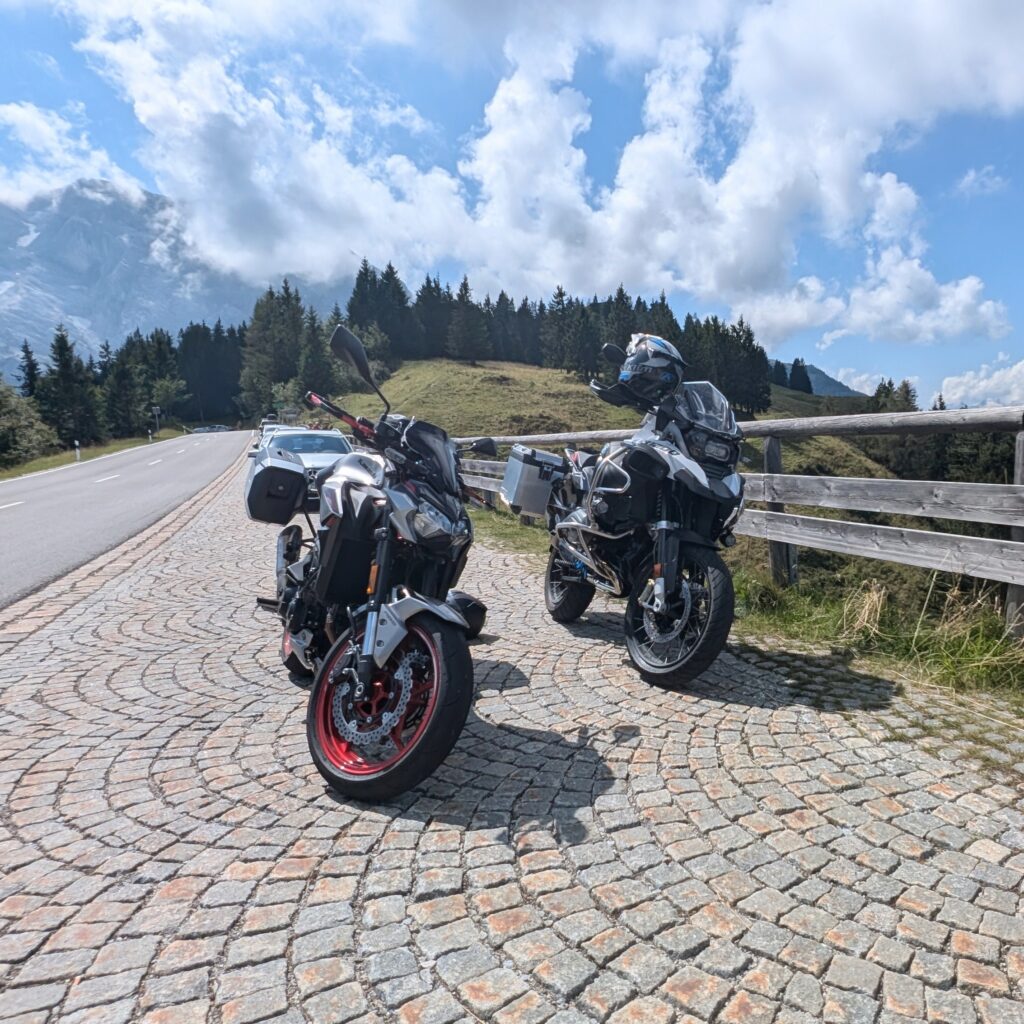 Zwei geparkte Motorräder (Kawasaki Z900 und BMW GS) auf einem Kopfsteinpflaster-Parkplatz in den Bergen neben einer Straße.