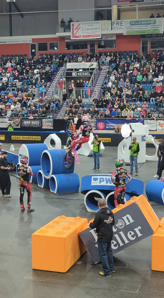 Motorrad-Trial-Fahrer in rot-schwarzer Montur springt bei einem Hallen-Wettbewerb über gestapelte blaue Röhren und orange Blöcke.