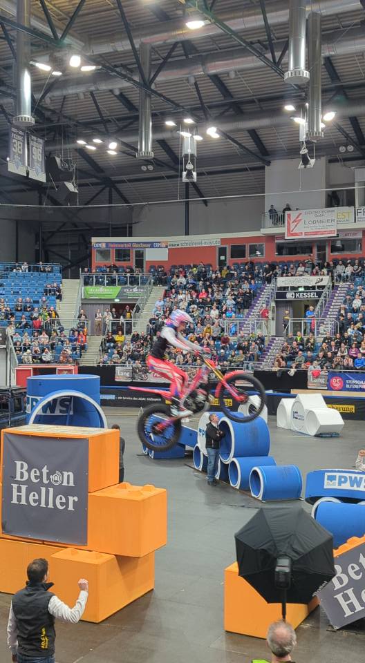 Motorrad-Trial-Fahrer in roter Kleidung springt in einer Sporthalle über blau-weiße Hindernisse und orangefarbene Beton Heller Blöcke.