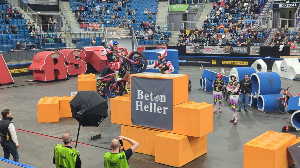 Motorrad-Trial-Wettbewerb in einer Arena mit Zuschauern, Fahrer auf orangefarbenen Blöcken mit "Beton Heller"-Logo.