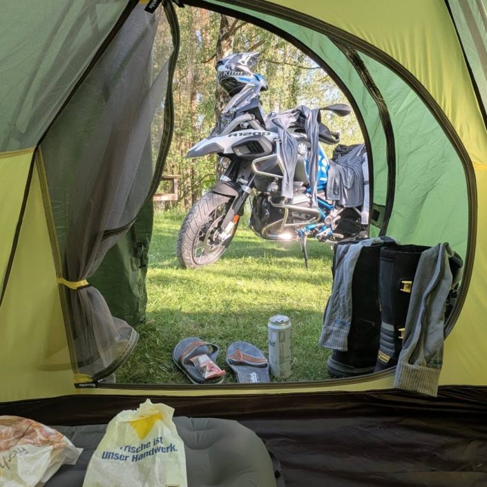 Blick aus einem gelb-grünen Zelt auf ein weiß-blaues BMW R 1200 GS Motorrad mit Helm im Grünen.