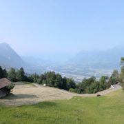 Blick von einer grünen Almwiese mit Scheune und Straße hinunter in ein weites Tal mit Fluss, Autobahn und Städten.