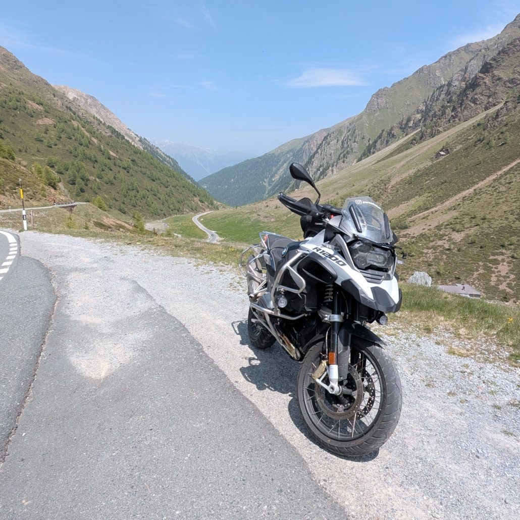 Silber-schwarzes BMW R 1200 GS Motorrad parkt auf Schotter neben einer Passstraße mit Blick in ein grünes, sonniges Hochgebirgstal.