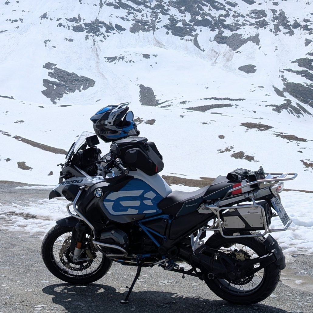 Weiße und blaue BMW R1200 GS Adventure mit Gepäck, Seitenkoffern und Helm vor einem schneebedeckten Berg.