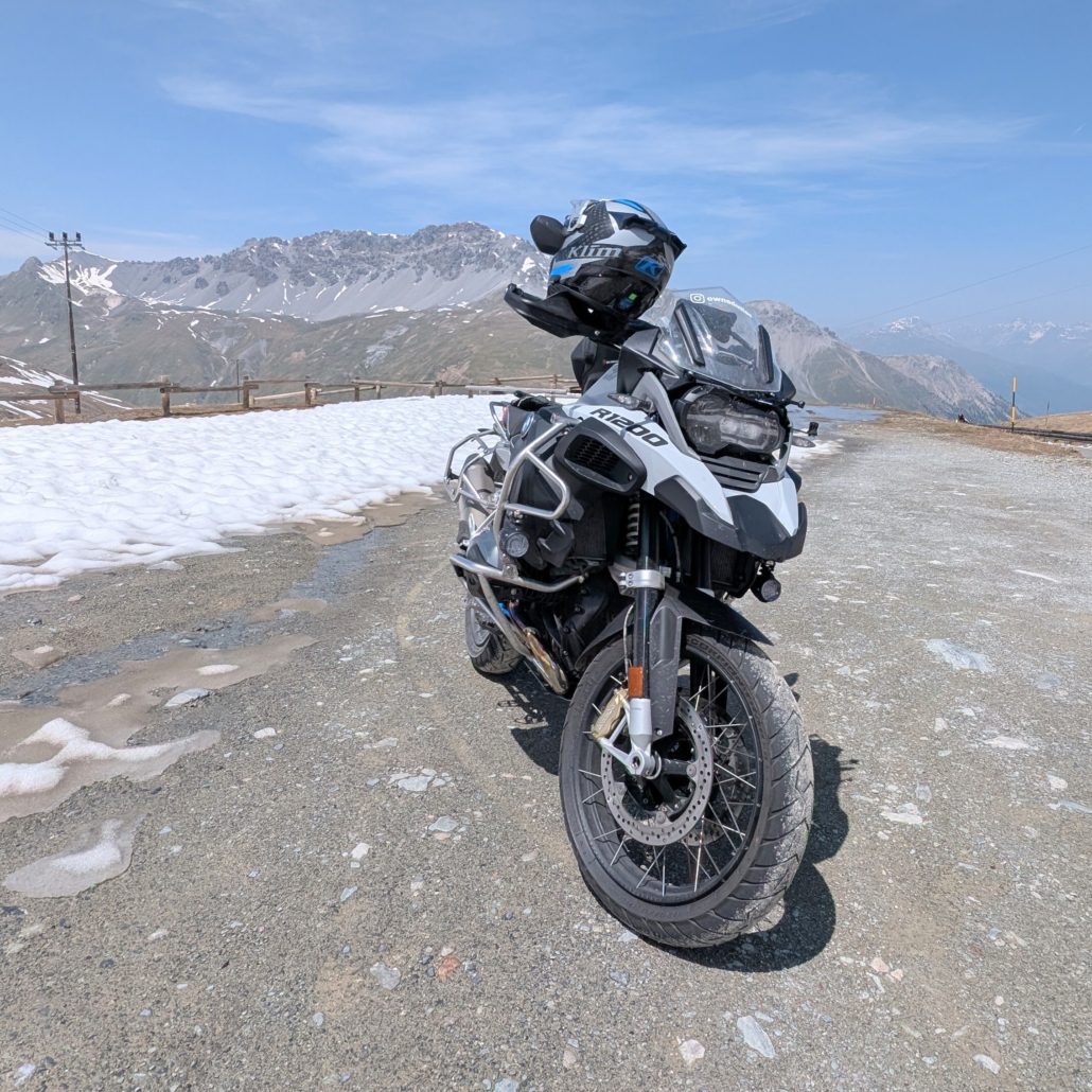 BMW R 1200 GS Adventure Motorrad mit Helm auf einem Schotterweg in den schneebedeckten Alpen unter blauem Himmel.