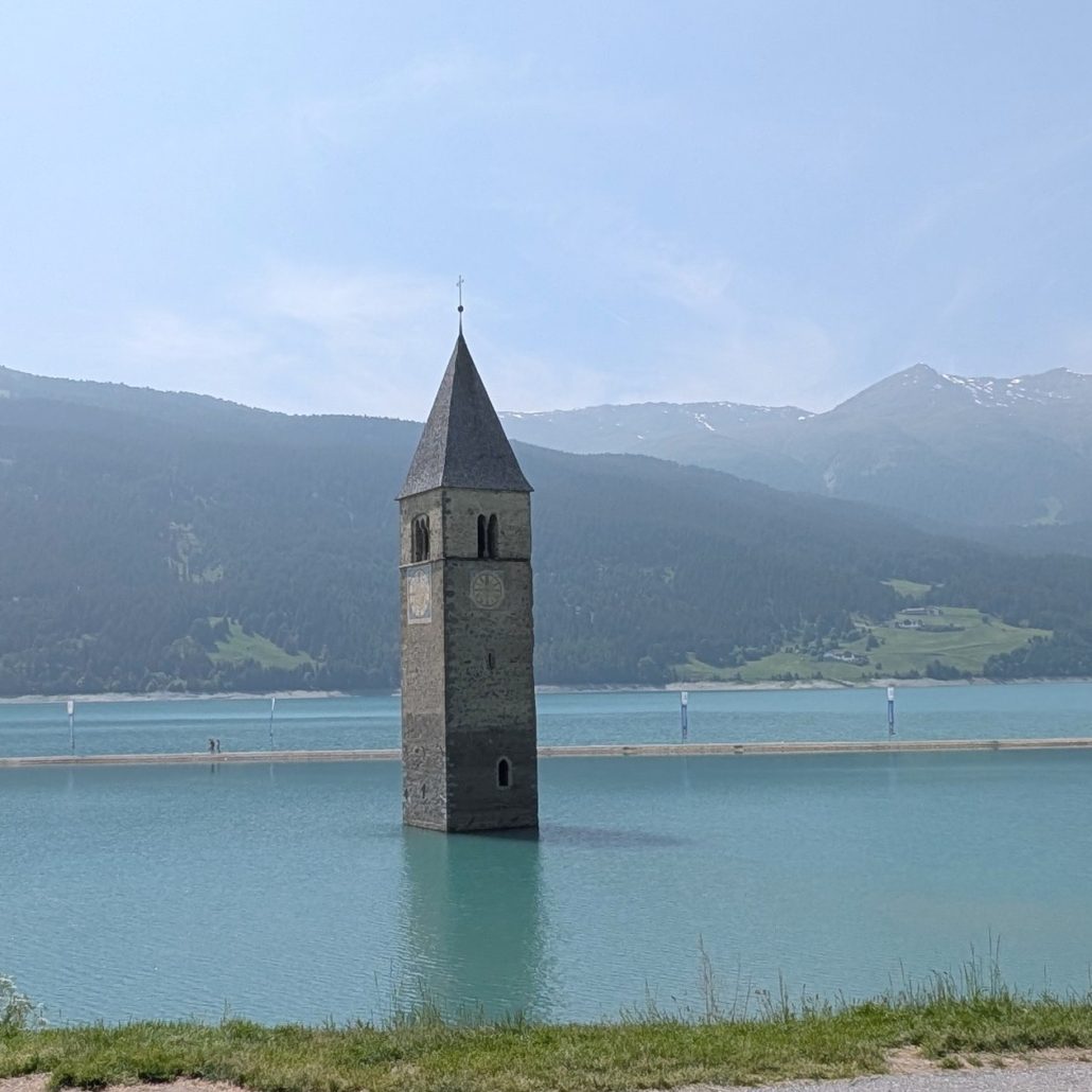 Unterwasser-Kirchturm von Graun im Reschensee (Südtirol), umgeben von türkisfarbenem Wasser und bewaldeten Bergen.