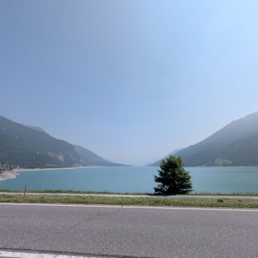 Stausee in einem Alpental mit bewaldeten Hängen und einem einzelnen Baum am Ufer.