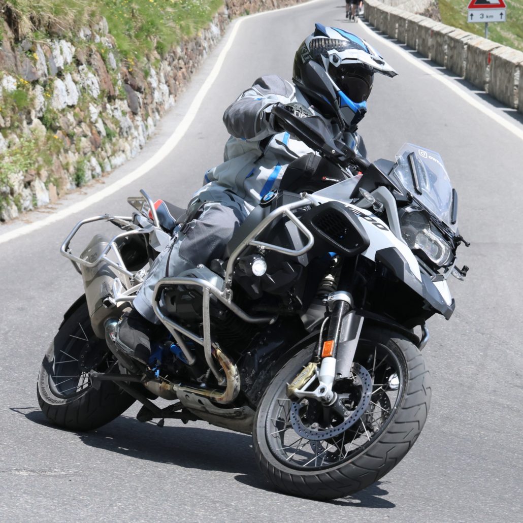 Motorradfahrer in grau-blauer Adventure-Kleidung fährt weiße BMW R 1200 GS mit Sturzbügeln auf einer Bergstraße.