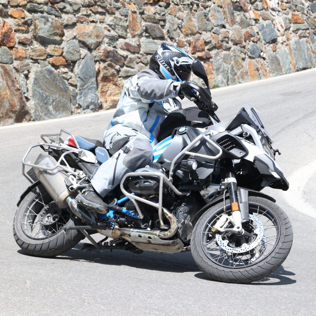 Motorradfahrer in grauer Adventure-Kleidung fährt eine weiße BMW R 1200 GS mit Sturzbügeln an einer Steinmauer.