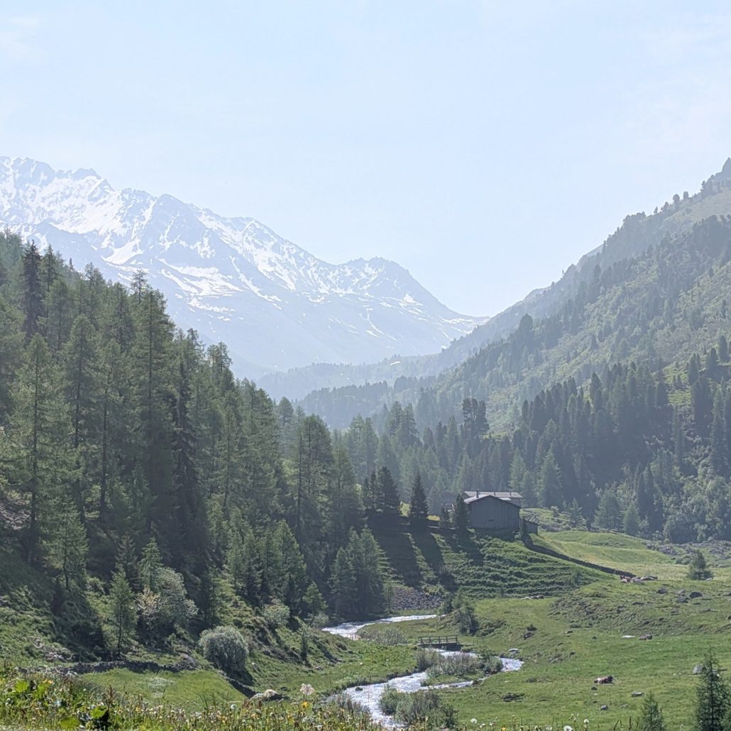 Straße am Bergpass neben einem See mit schnee-bedeckten Gipfeln im Hintergrund, im Vordergrund ein Motorrad-Helm.