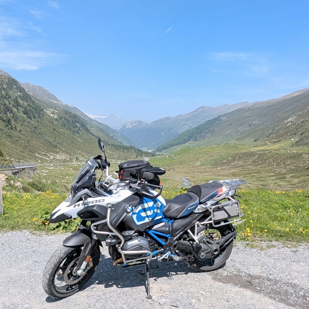 Weiße BMW R 1200 GS Adventure auf Schotterweg in einem grünen Hochgebirgstal mit weiten Wiesen und Bergen.
