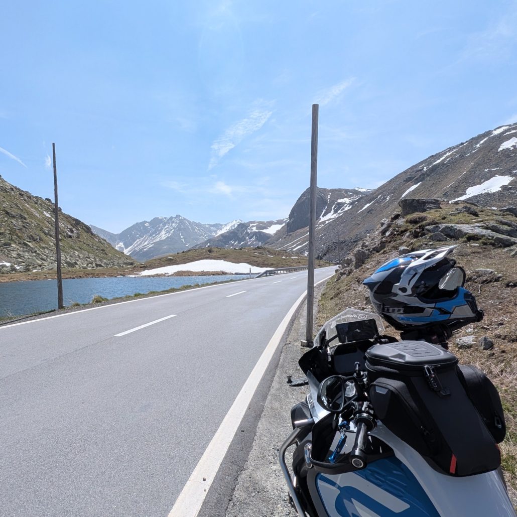 Motorrad mit Helm und Tankrucksack am Straßenrand eines Bergpasses, links ein See, rechts schneebedeckte Gipfel.