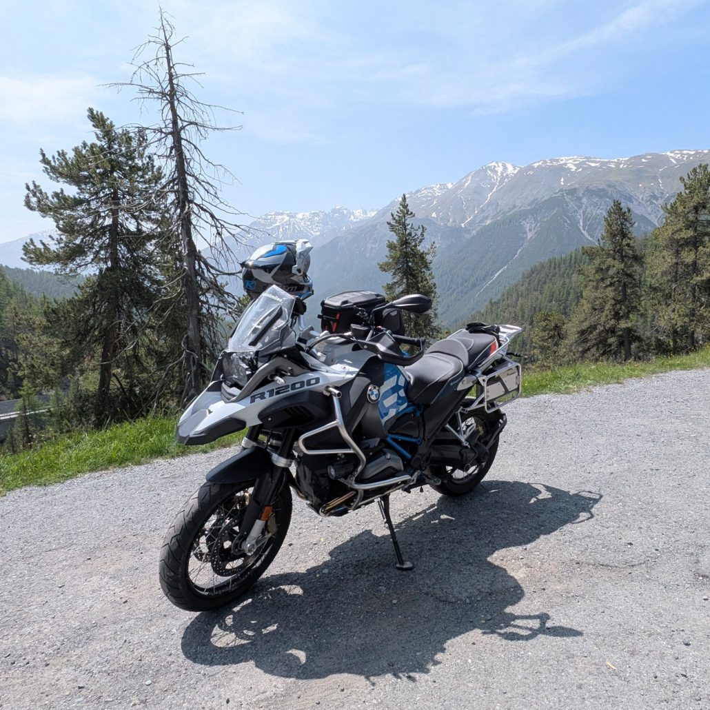 Silber-schwarzes BMW R 1200 GS Adventure Motorrad auf Schotterstraße vor bewaldeter Berglandschaft mit Schnee.